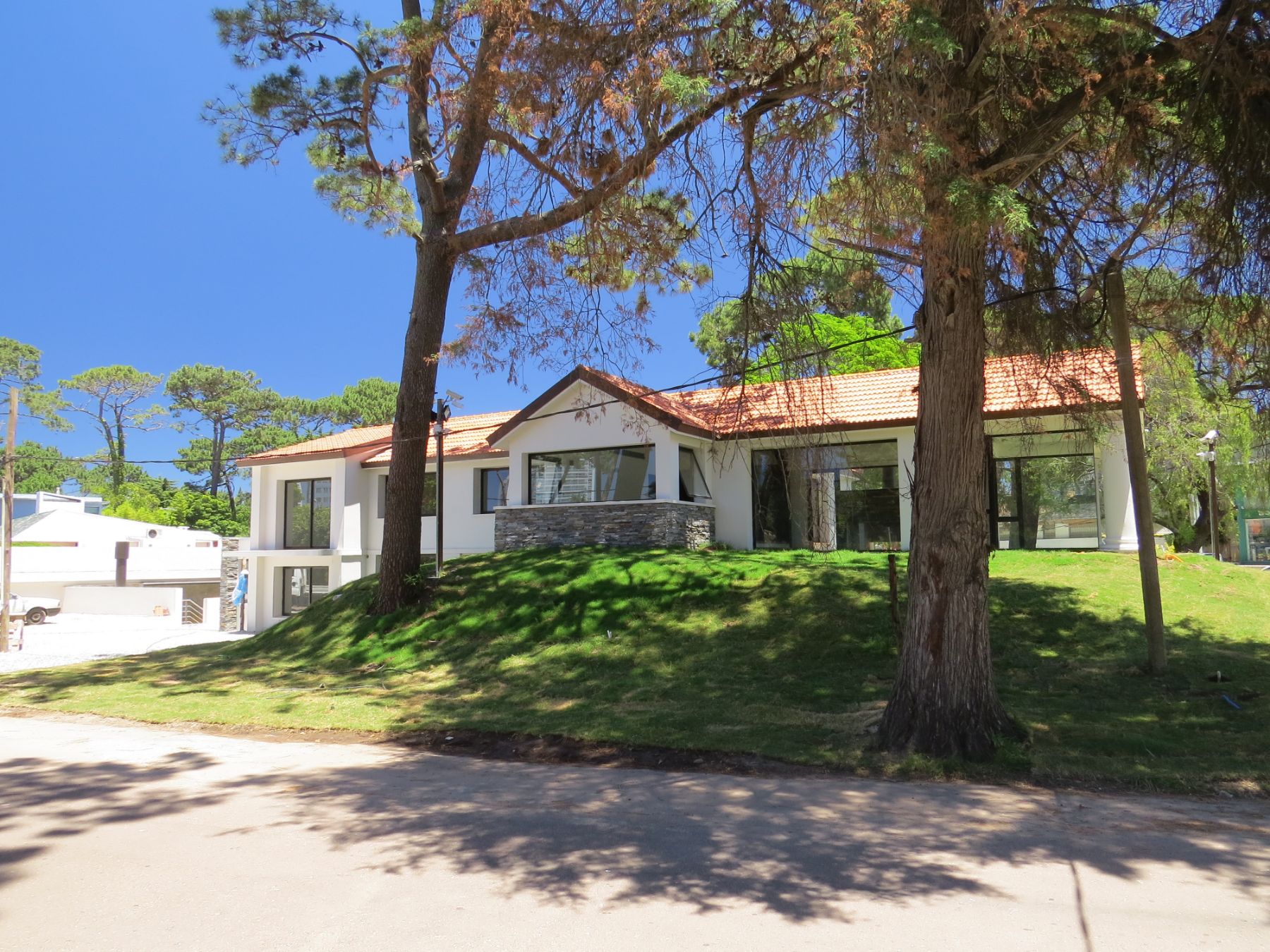 En la fotografía se ve la entrada latteral a la clínica de punta del este de blue cross. La misma posee un saliente en el centro de la construcción, techado a dos aguas con tejado y a partir de él nacen las dos alas de la clínica, todo esto sobre una pequeña colina.
